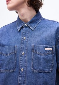 Άτομο που φοράει μπλε πουκάμισο από τζιν με κουμπιά και δύο μπροστινές τσέπες στο στήθος, από τις οποίες η μία φέρει ετικέτα με την ένδειξη «Calvin Klein».