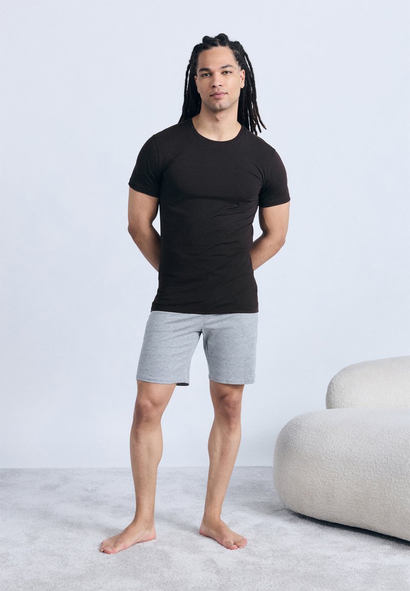 Νέος άνδρας με μακριές πλεξούδες, φορώντας μαύρο t-shirt και γκρι σορτς, όρθιος ξυπόλυτος σε χαλί δίπλα σε καμπύλο λευκό καναπέ.