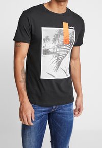 Sort bomulds t-shirt med et rektangulært grafisk motiv af en strandscene med palmer, suppleret med en lys orange detalje nær halsudskæringen.