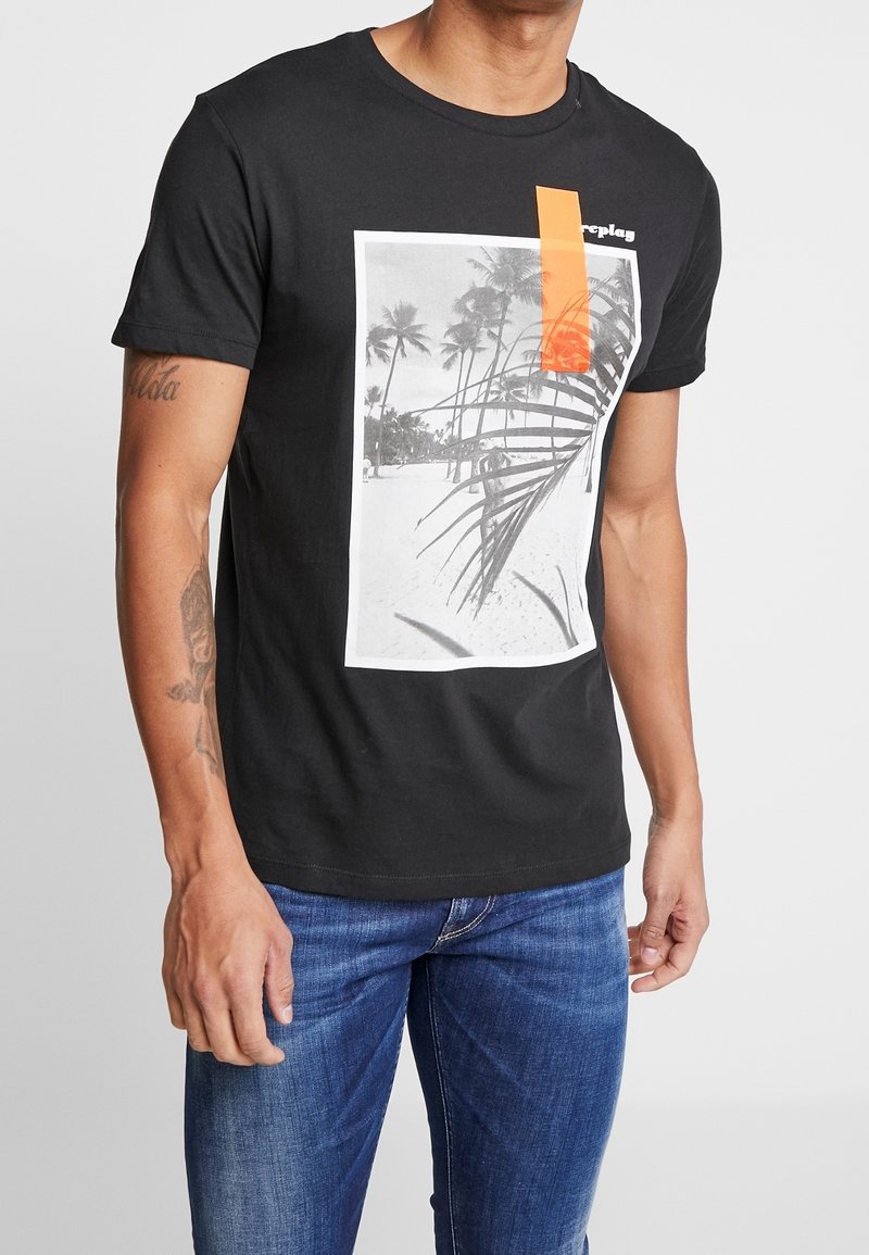 Sort bomulds t-shirt med et rektangulært grafisk motiv af en strandscene med palmer, suppleret med en lys orange detalje nær halsudskæringen.