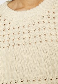 Pletený svetr v krémové barvě, se strukturovaným vzorem otevřených a uzavřených stehů, žebrovaným límcem a lemem a měkkým, vrstveným vzhledem.