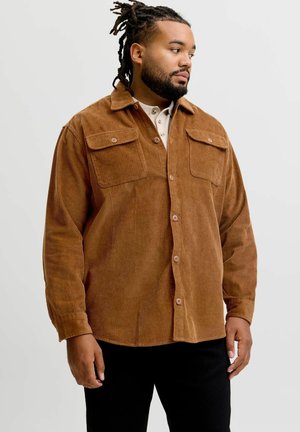 Mand med dreadlocks iført en brun kordfløjls-skjorte med knapper over en lys beige henley, stående mod en ensfarvet lys baggrund.