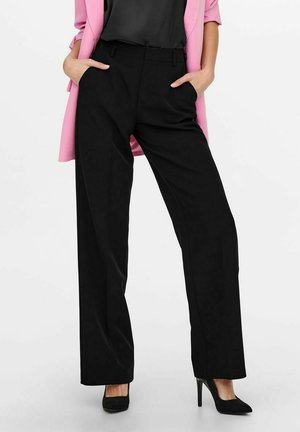Pantalon noir sur mesure à jambe droite, en tissu léger, avec poches latérales. Porté avec un blazer rose et des escarpins noirs.