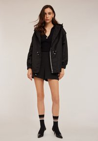 Svart nylonjacka med huva, elastiska ärmslut och justerbara dragsko. Kombinerad med en svart topp, shorts och knähöga strumpor med ränder.