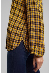 Camicia a maniche lunghe giallo e nera a scacchi con orlo arrotondato e spacchi laterali, realizzata in tessuto intrecciato, con dettagli arricciati sulle maniche.
