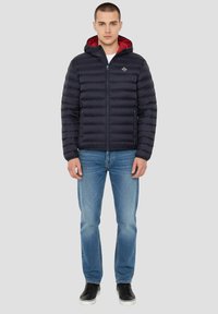 Doudoune bleu marine avec une doublure intérieure rouge, fermeture éclair, capuche et deux poches latérales, portée avec un jean bleu et des baskets noires.