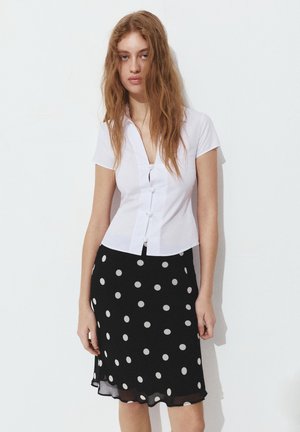 Junge Frau mit langem Haar, die eine weiße Bluse mit kurzen Ärmeln und einen schwarzen Rock mit Punkten trägt, steht vor einer schlichten weißen Wand.