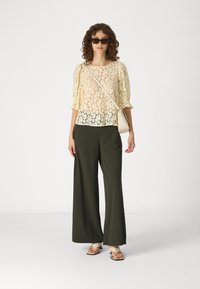 Krajkový top v bledě žluté barvě, s nabíranými rukávy; volné olivově zelené kalhoty; béžové sandály se strapci; jednoduchá krémová crossbody taška; sluneční brýle.