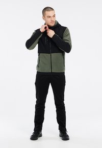 Fleecejacke in Olivgrün und Schwarz, mit durchgehendem Reißverschluss, Kapuze und Reißverschlusstaschen. Schlanker Schnitt mit kontrastierenden Stoffbereichen.