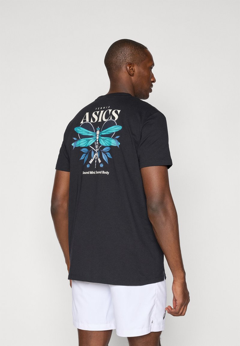 Mann im Seitenprofil, trägt ein schwarzes ASICS-Tennis-T-Shirt mit blauem Libellenmotiv und weiße Shorts, vor weißem Hintergrund.