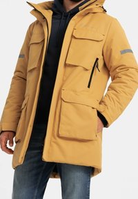 Parka aislante amarilla con cierre de cremallera, múltiples bolsillos con solapa y cremallera, y detalles reflectantes. Cuenta con capucha y un corte relajado.