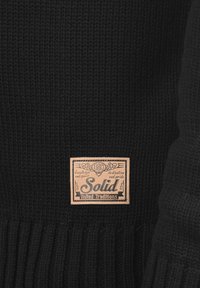 Schwarzer Strickpullover mit geripptem Saum und einem beige Etikett mit den Aufschriften "Solid" und "United Traditions", auf einem dunklen Hintergrund.