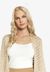 Beige virkad kardiagan med öppen design, bärs över en vit linne. Långt, vågigt blont hår och färgglada hängande örhängen.