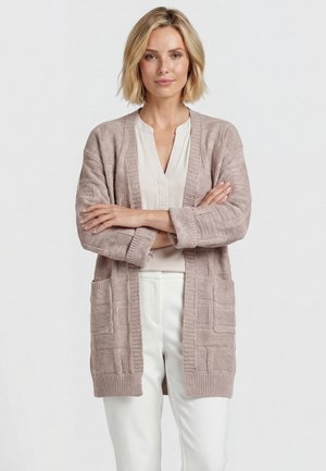 Vrouw met blond haar die een beige gestructureerde cardigan draagt over een witte blouse en witte broek, staand met gekruiste armen tegen een lichte achtergrond.
