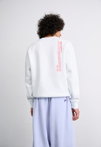 Sweatshirt branco de mangas longas, apresentando texto impresso em vermelho nas costas. Combinado com calças largas de um tom roxo claro e detalhe de logo.