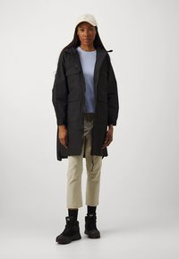 Svart vanntett parkas med glidelås foran, to brystlommer og justerbare mansjetter, båret over en lys blå topp og beige bukser. Svarte støvler.