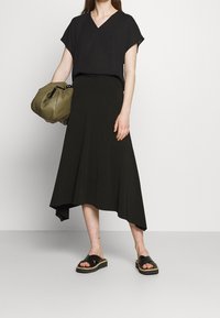 Haut noir à col en V et jupe noire asymétrique. Modèle tenant un sac vert olive. Sandales à plateforme noires avec larges brides. Textures lisses.