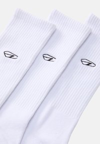 Bijele rebraste čarape s crnim logotipom sprijeda. Tkanina izgleda mekano s teksturiranim završetkom. Prikazane su tri para.