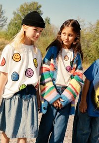 Två barn som bär t-shirts med grafiska tryck med leende ansikten. En flicka har på sig en flerfärgad randig kofta, och båda har jeans eller denimbyxor.