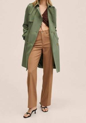 Kvinde iført en olivengrøn trenchcoat, brune høj-taljede lige bukser, en mørkebrun crop top og sorte sandaler med hæl.