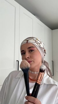 Två sminkborstar hålls upp, en med en fluffig huvud med blå spets och den andra med ett grått och vitt huvud, båda med svarta handtag. En vit skjorta och ett mönstrat huvudscarf bärs.