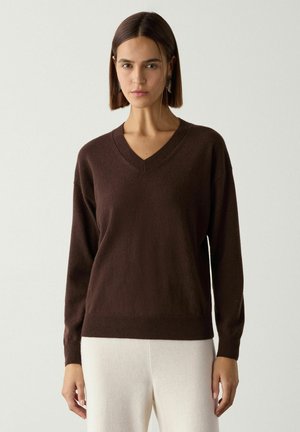 Kvinde med glat brunt hår iført en mørkebrun V-hals sweater og lyse cremefarvede bukser mod en ensfarvet lys baggrund.