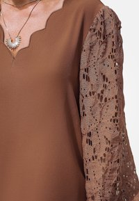 Braune Bluse mit einem gewellten Ausschnitt und halbtransparenten Spitzenärmeln mit Blattmuster. Glatte Stoffoberfläche mit leicht glänzendem Finish.