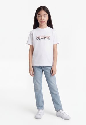 Ragazza giovane in piedi, indossa una T-shirt bianca con grafica, jeans azzurri chiari e sneakers bianche su sfondo bianco uniforme.