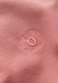 Rosa tyg med en strukturerad väv som har en broderad logotyp, en krona och ett lagerdesign i en något mörkare rosa nyans.