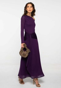 Mujer vestida con un largo vestido morado de un solo hombro, con la cintura fruncida, llevando un bolso marrón de estampado y usando sandalias de tacón alto doradas.