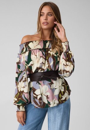 Donna con una camicetta floreale scollata con maniche a sbuffo e cintura scura, abbinata a jeans azzurri su sfondo neutro.