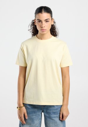 Jonge vrouw met krullend donker haar in een paardenstaart, gekleed in een lichtgele T-shirt met korte mouwen en een blauwe spijkerbroek, tegen een effen achtergrond.