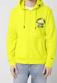 Felpa gialla con zip, coulisse, tasche frontali e testo nero stampato sul petto. Tessuto morbido e polsini a costine.