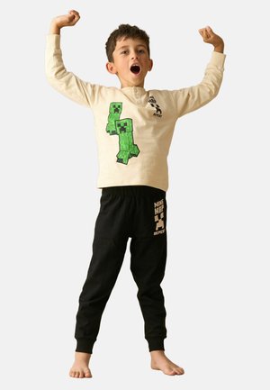 Garçon en pyjama sur le thème de Minecraft, levant les bras avec la bouche ouverte, debout pieds nus sur un fond uni.