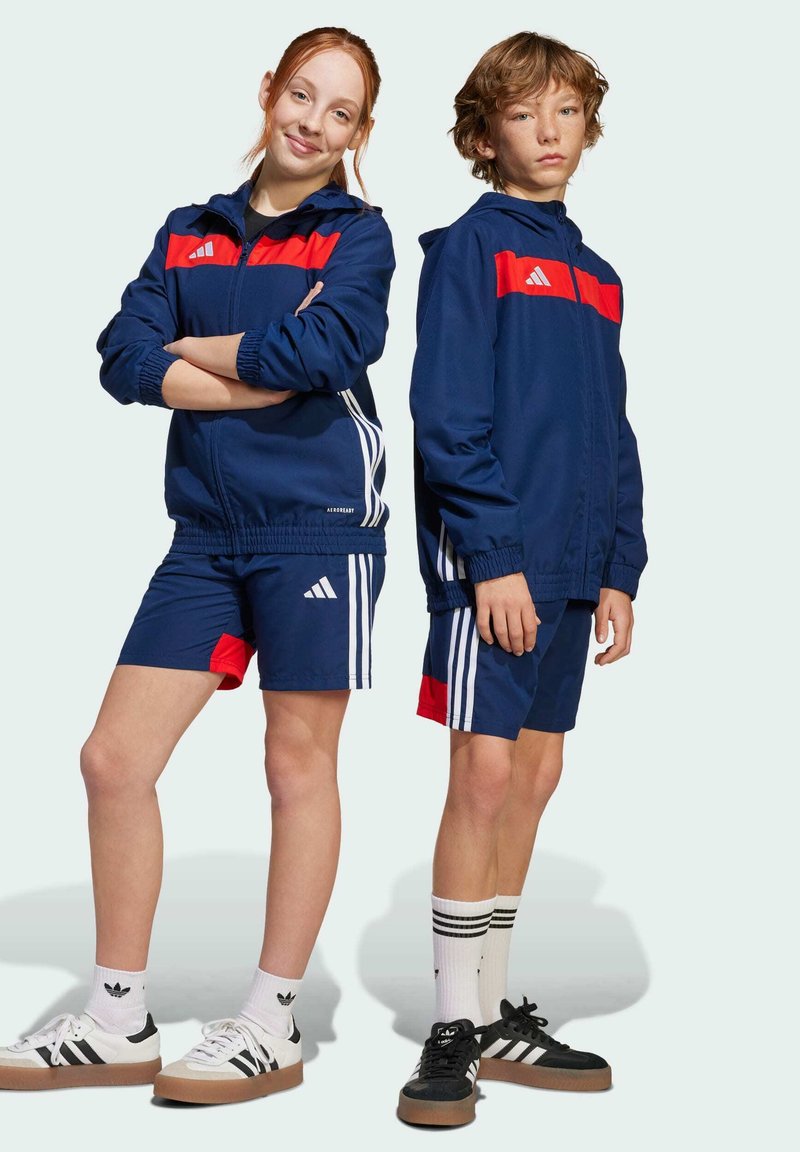 Navy sportswear set featuring a jacket with red accents and striped sides, paired with shorts. Both wear sneakers and striped socks.