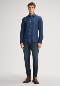 Camicia button-up in denim blu scuro con colletto, maniche lunghe e bottoni bianchi. Abbinata a jeans in denim scuro e stivali in pelle marrone.