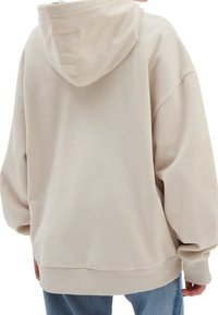 Beige mikina s kapucí a volným střihem, s žebrovanými manžetami a lemem. Měkká, texturovaná tkanina viditelná na zádech, s neformálním siluetou.