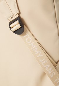 Rucsac din material bej, cu curea ajustabilă, având inscripția "TOMMY JEANS" în text alb, evidențiind o suprafață texturată și o cataramă rezistentă.