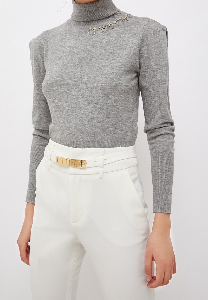 Pull gris à col roulé avec texture côtelée, orné de décorations près de l'épaule, associé à un pantalon blanc et une ceinture dorée.