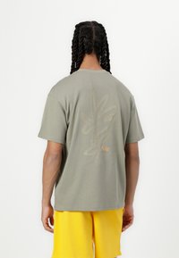 Olive zaļas kokvilnas T-krekls ar smalku teksturētu lapu rakstu gaiši brūnā krāsā un mazu oranžu Nike logo aizmugurē.
