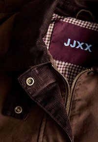 Brown corduroy jacket collar with brass snap buttons, zipper, and checked inner lining showing a maroon label with white "JJXX" text.