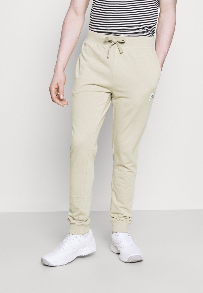 Ljusbeige joggers med dragsko i midjan, ribbade muddar och en liten logotyp-patch på sidan, parat med vita sportskor.