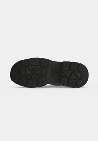 Semelle de chaussure noire avec des crampons profonds, des motifs géométriques et le logo Guess embossé en forme triangulaire au centre.