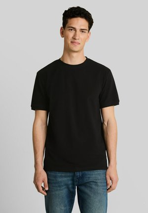 Junger Mann mit dunklem lockigem Haar, der ein schlichtes schwarzes T-Shirt mit kurzen Ärmeln und blaue Jeans trägt, steht vor einem grauen Hintergrund.