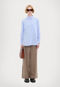 Camicia azzurra chiara con colletto e tasca, abbinata a pantaloni larghi marroni, scarpe nere con suola spessa e una borsa marrone.