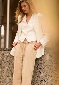 Weißer Leinenblazer über einer kurzen weißen Bluse, kombiniert mit beigen Bundhosen. Die glatten Texturen und neutralen Töne werden betont.