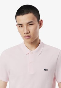 Ljust rosa polo skjorta med klassisk krage, treknapps placket, texturerad tyg och en liten broderad logotyp på vänster bröst.