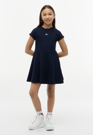 Mladé dievča stojí s rukami za chrbtom, oblečené v námornícky modré šaty s krátkym rukávom a biele vysoké tenisky s bielymi ponožkami.