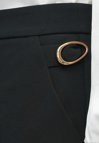 Μαύρο ύφασμα με ομαλή υφή που διαθέτει διακοσμητικό χρυσό βρόχο στο πλάι, προσαρτημένο σε λεπτομέρεια τσέπης.