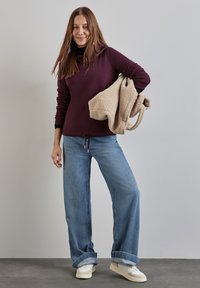 Top lungo bordeaux con collo a cascata, abbinato a jeans a gamba larga blu chiaro e scarpe da ginnastica bianche; tiene in mano una borsa beige morbida.